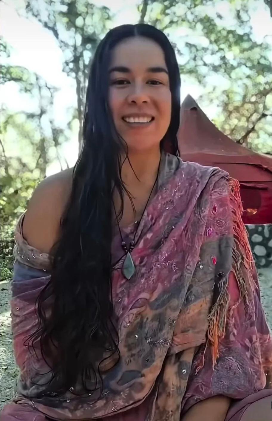 Smiling woman with long dark hair sits outdoors in a pink patterned wrap, with a red tent behind her and trees in the background.
