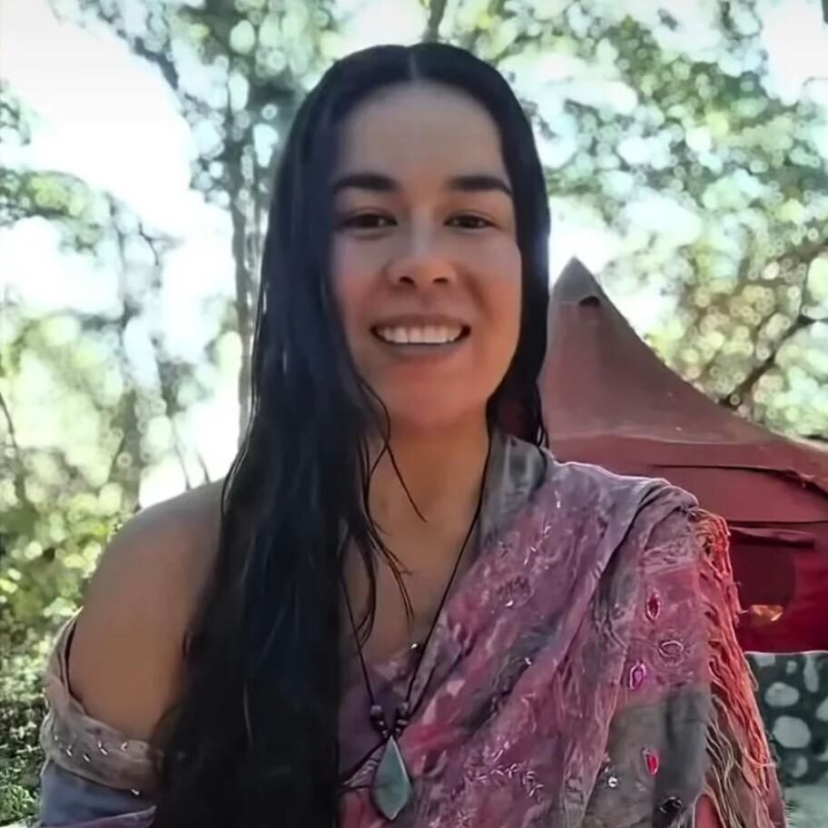 Woman with long dark hair smiling outdoors, wearing a pink patterned shawl and a pendant, with a tent in the background.