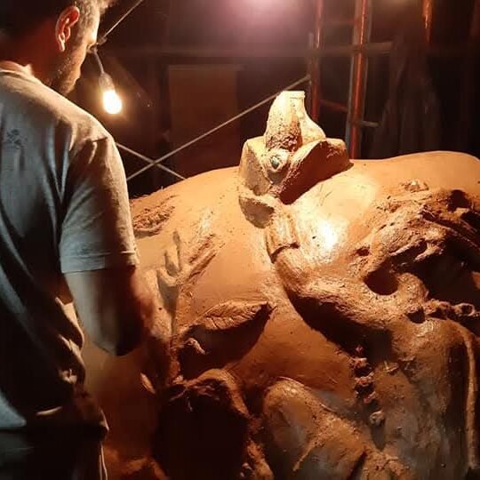 Man in a gray t-shirt sculpting a large clay statue under a single hanging bulb in a dim workshop.